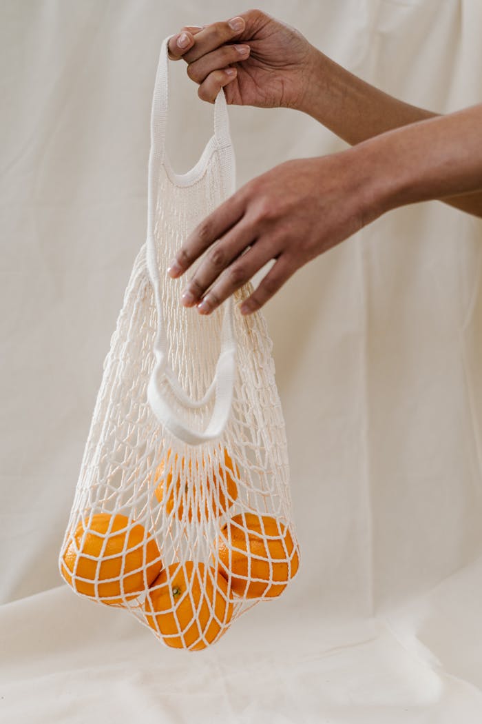 Hand holding a reusable mesh bag filled with fresh oranges against a neutral background.