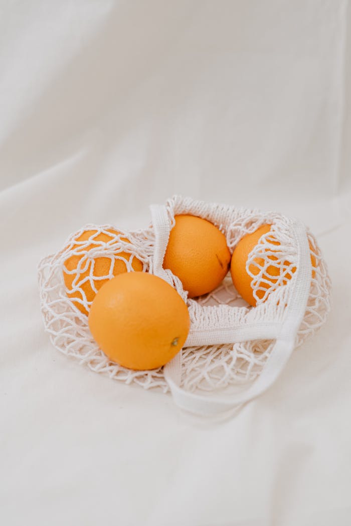Four ripe oranges in an eco-friendly mesh bag on a clean white background.
